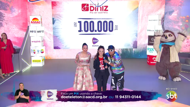 Da esquerda para a direita: Silvia Abravanel, Claudia Valerio e Liminha estão no palco do Teleton, com o telão atrás exibindo o valor da doação das Óticas Diniz para o Teleton. O número cem mil é exibido no telão.