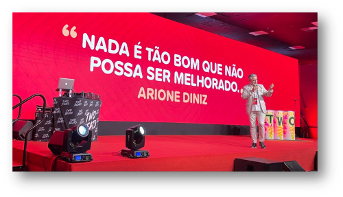 Arione Diniz está no palco, em frente a um telão que exibe a frase: "Nada é tão bom que não possa ser melhorado".