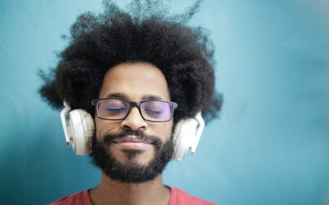 Homem aparentemente de Home Office, sentado em frente do computador usando óculos de grau e fones de ouvido. Sua expressão é de tranquilidade.